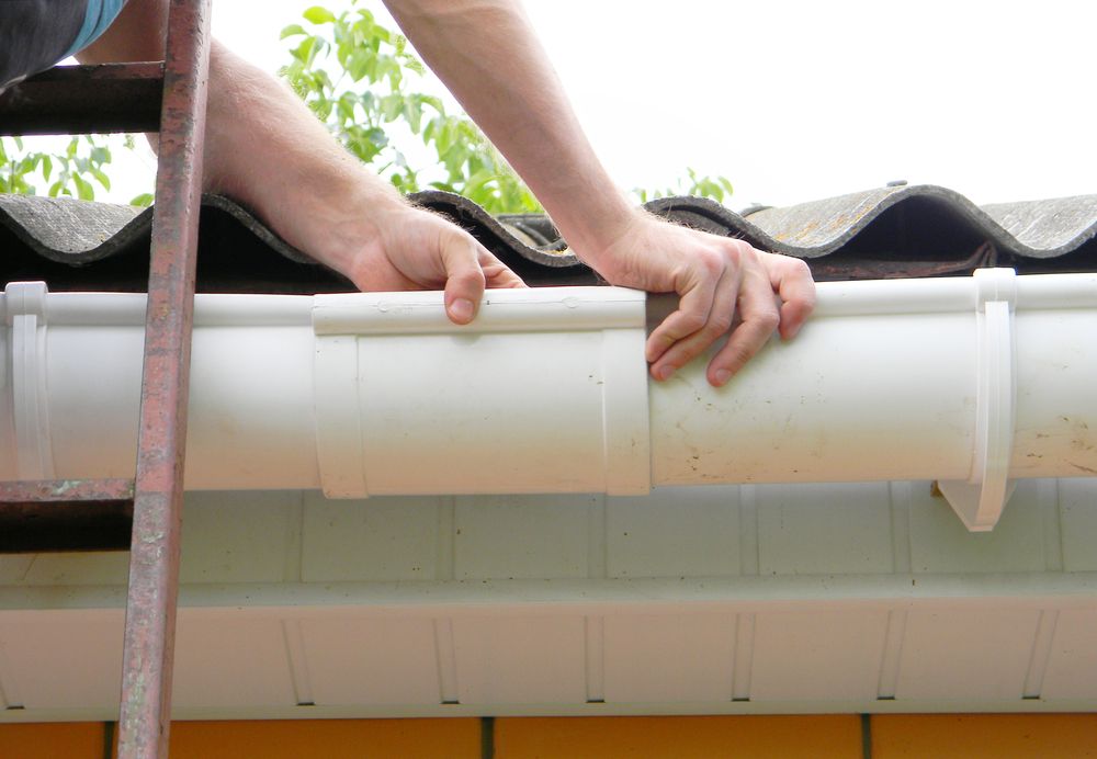 A Person is Standing on a Ladder Fixing a Gutter on a Roof — Norwest Roof Restorations In Blue Bay, NSW